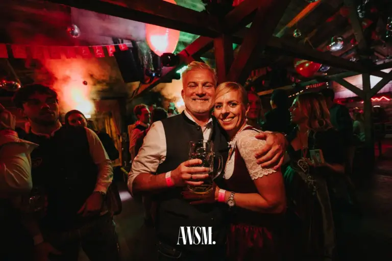 Mann und Frau in Tracht posieren Arm in Arm mit Bierkrug unter dem Holzdach mit Ballons und Wimpelgirlande – AWSM. GOES KAISER WIESN, 27.09.2026