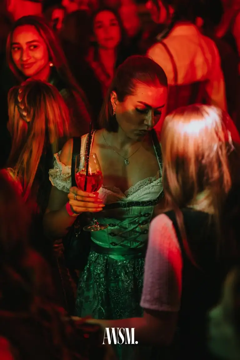 Frau im grün-weißen Dirndl hält ein Glas mit rotem Getränk und Strohhalm, ringsum Gäste im roten Licht – AWSM. GOES KAISER WIESN, 27.09.2026