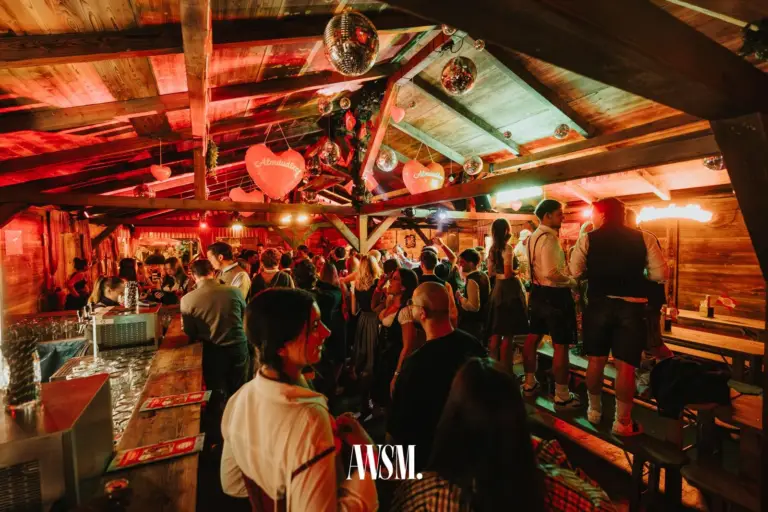 Blick unter das Holzdach auf der Kaiserwiese im Prater: Bar links, Gäste in Tracht, Discokugeln und Herzdeko – AWSM. GOES KAISER WIESN, 27.09.2026