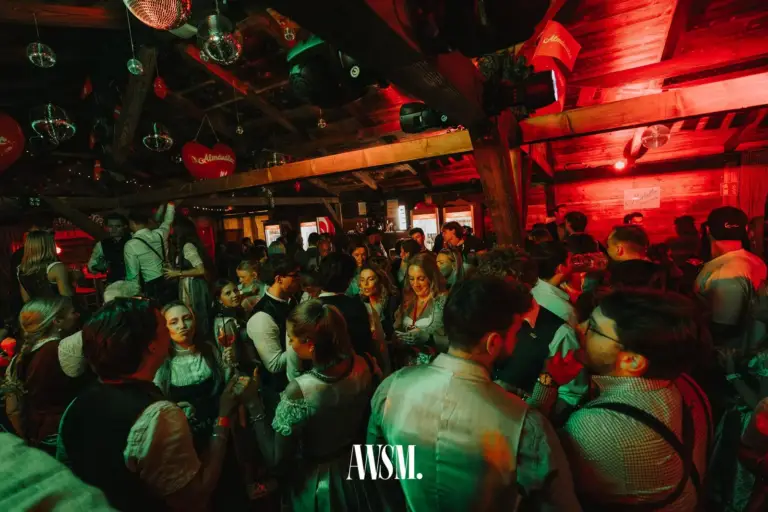 Dicht gedrängte Gäste in Tracht unter Holzbalken mit Discokugeln, Herzballons und rotem Licht – AWSM. GOES KAISER WIESN, 27.09.2026