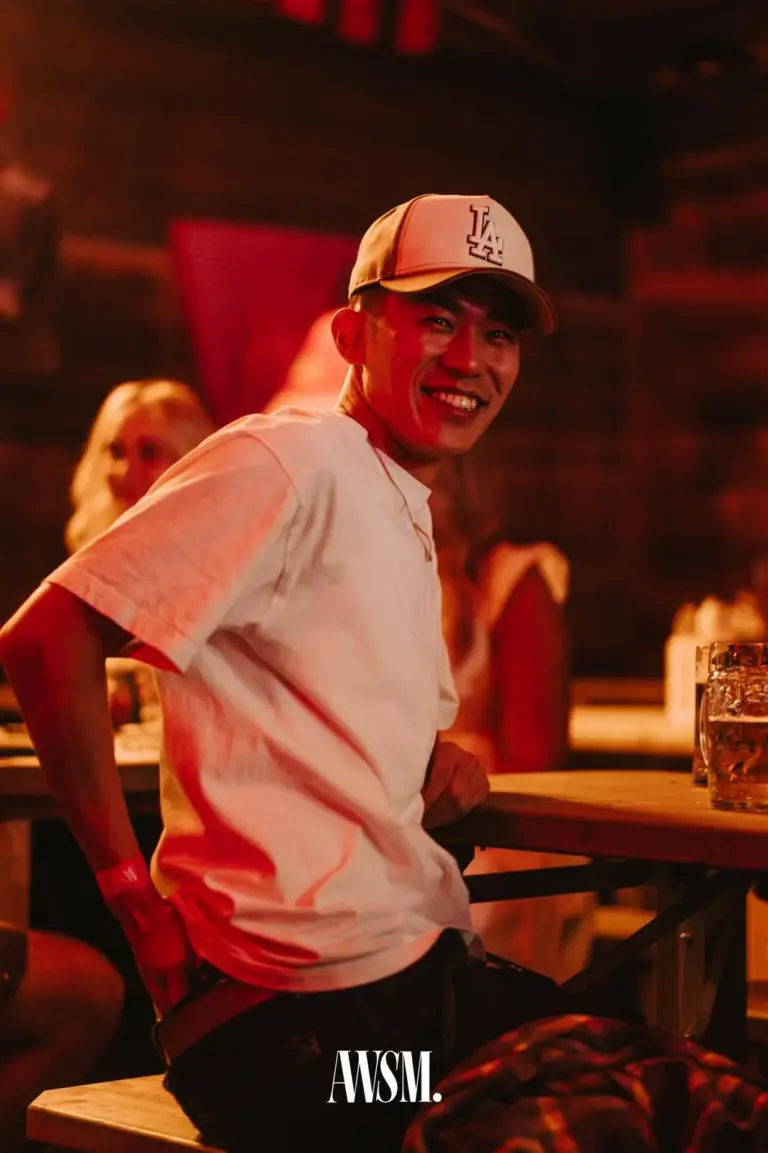 Mann in weißem T-Shirt und Kappe lacht auf der Bierbank am Tisch mit Bierkrug, rotes Licht – AWSM. GOES KAISER WIESN, 27.09.2026