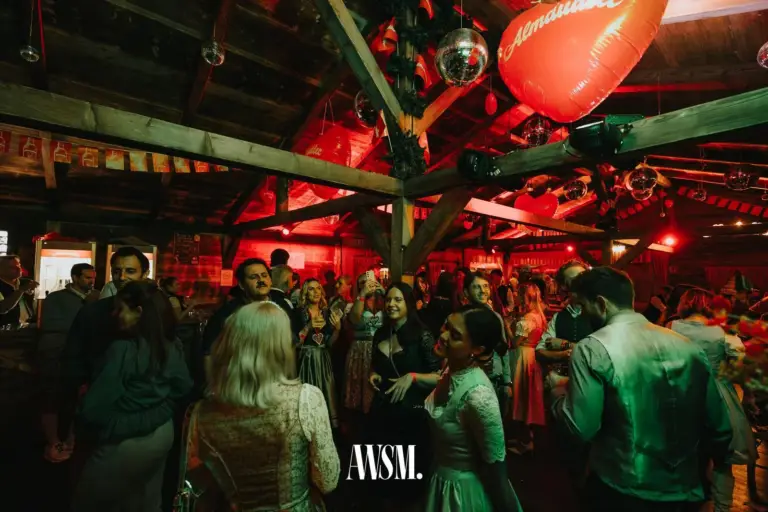 Gäste in Tracht tanzen im grünen Licht unter Holzbalken, darüber ein großer Herzballon und Discokugeln – AWSM. GOES KAISER WIESN, 27.09.2026