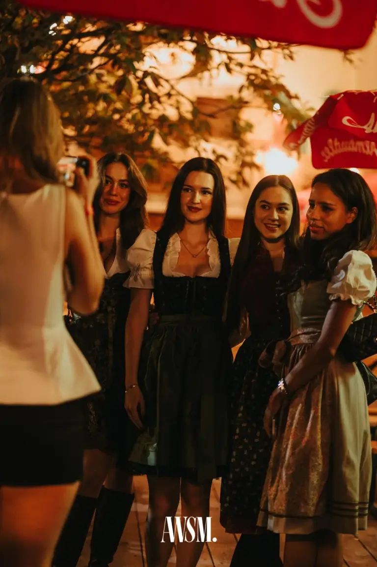 Vier Frauen im Dirndl posieren im Freien unter einem Baum und Schirmen, eine Frau fotografiert sie mit dem Handy – AWSM. GOES KAISER WIESN, 27.09.2026