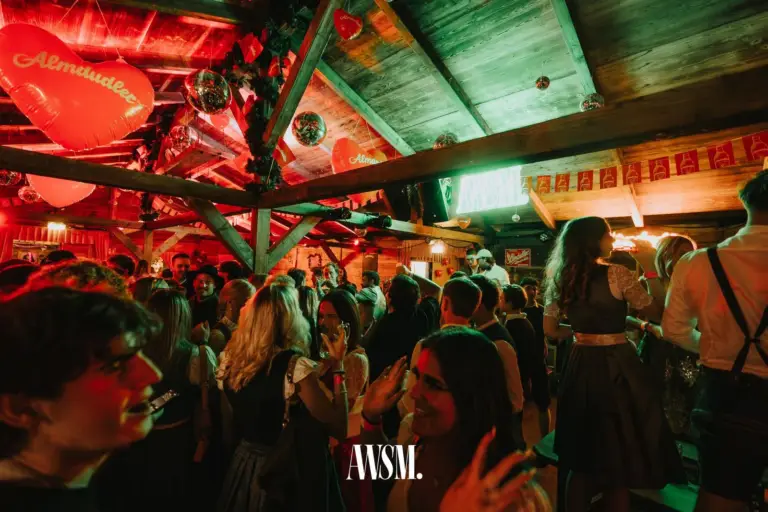 Feiernde Menge unter dem Holzdach mit Herzballons, Discokugeln und Wimpeln, vorne Gäste im Dirndl – AWSM. GOES KAISER WIESN, 27.09.2026