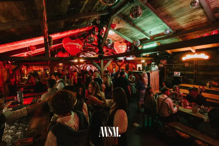 Weiter Blick unter das Holzdach mit Bar, Bierbänken und Herzdeko, dazwischen Gäste in Tracht – AWSM. GOES KAISER WIESN, 27.09.2026