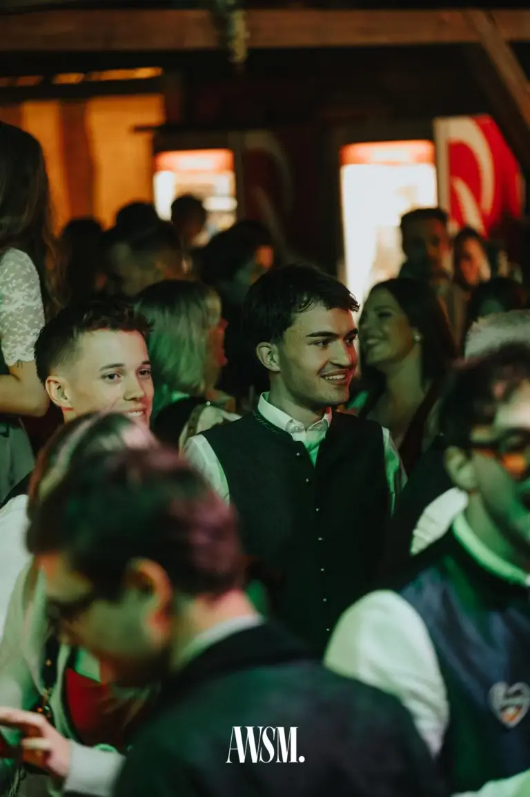 Männer in Trachtengilet und weißem Hemd lächeln in der Menge, warmes und grünes Licht – AWSM. GOES KAISER WIESN, 27.09.2026
