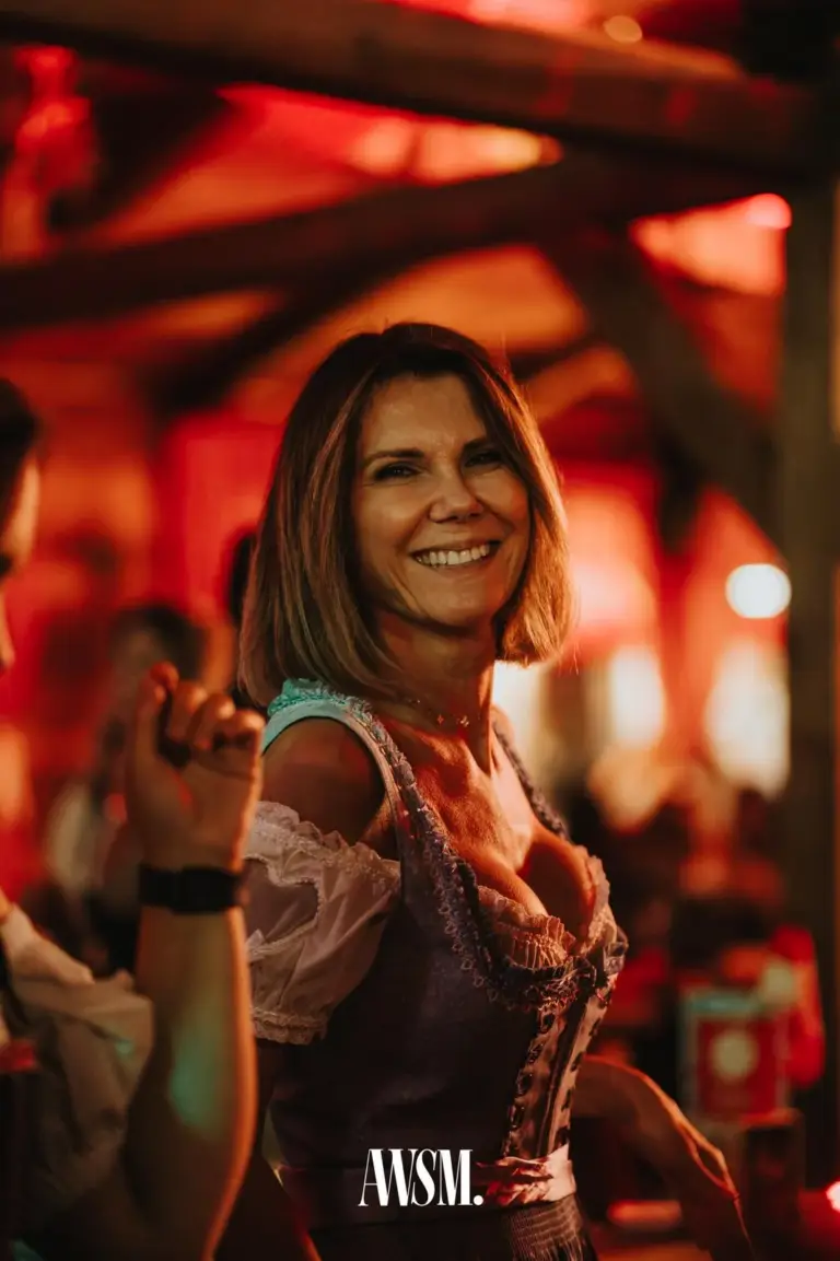 Frau im Dirndl mit Bob-Frisur lächelt in die Kamera, dahinter rot beleuchtete Holzbalken – AWSM. GOES KAISER WIESN, 27.09.2026