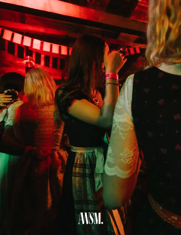 Gäste im Dirndl von hinten, eine Frau trinkt, darüber Wimpelgirlande und rot beleuchtete Holzbalken – AWSM. GOES KAISER WIESN, 27.09.2026