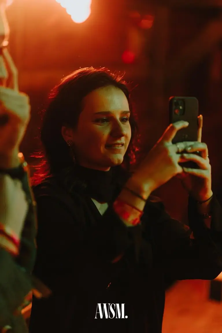 Frau in dunkler Kleidung filmt mit dem Handy, warmes rotes Licht im Hintergrund – AWSM. GOES KAISER WIESN, 27.09.2026