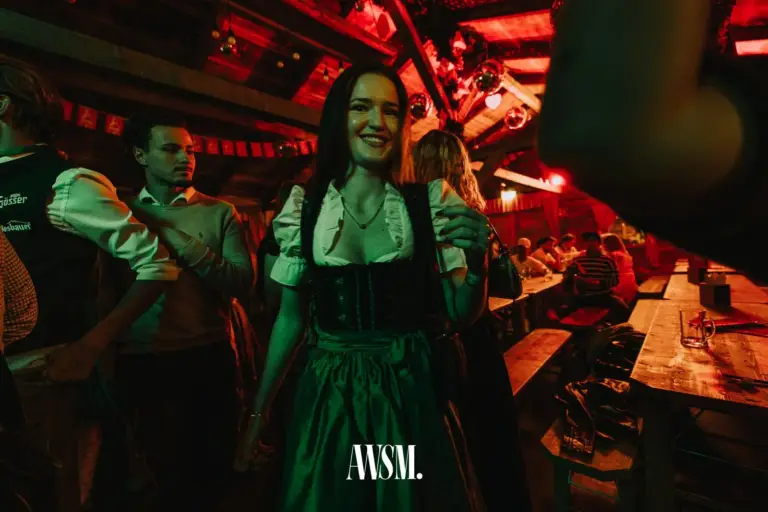Frau im grünen Dirndl lächelt zwischen Gästen und Bierbänken, rot beleuchtetes Holzdach – AWSM. GOES KAISER WIESN, 27.09.2026