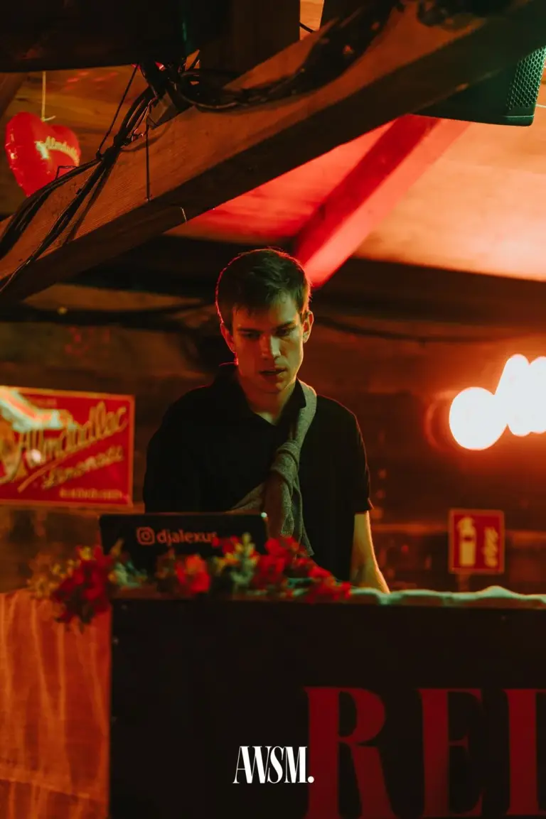 DJ am Pult mit Laptop und roten Blumen, darüber Holzbalken, Herzballon und rotes Licht – AWSM. GOES KAISER WIESN, 27.09.2026